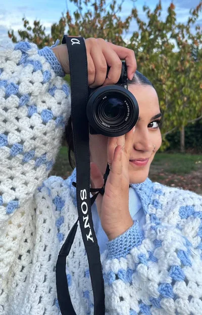 Maeva tient son appareil photo devant un œil, en extérieur, vêtue d’un gilet bleu et blanc